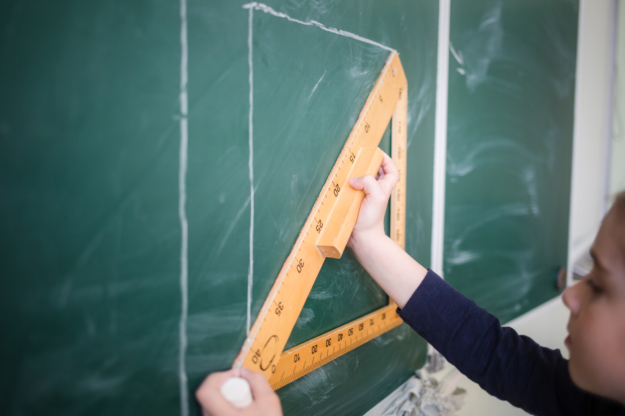 child draws line with chalk using triangle ruler on blackboard in school classroom