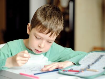cute and smiling boy in a green t-shirt draws with colored pencils while sitting at the table