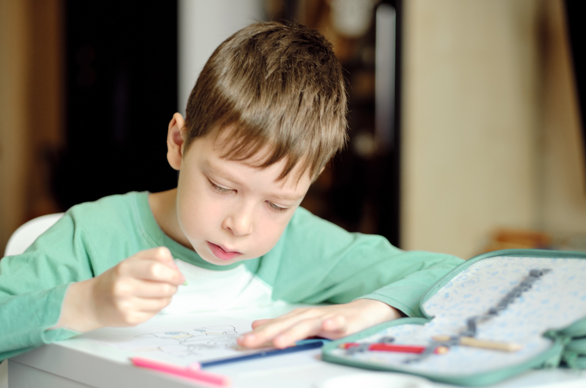 cute and smiling boy in a green t-shirt draws with colored pencils while sitting at the table