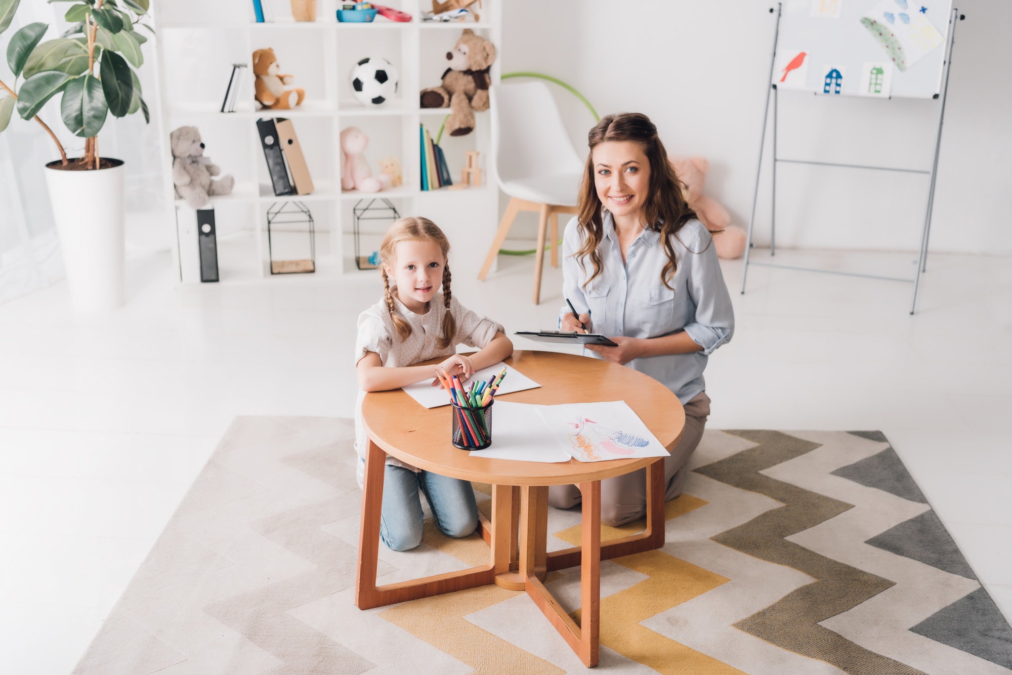 happy psychologist with clipboard sitting near little child while she drawing with color pencils and