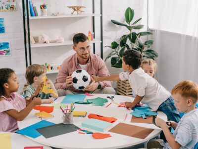 male teacher with soccer ball and multiracial preschoolers sitting at table with colorful papers in