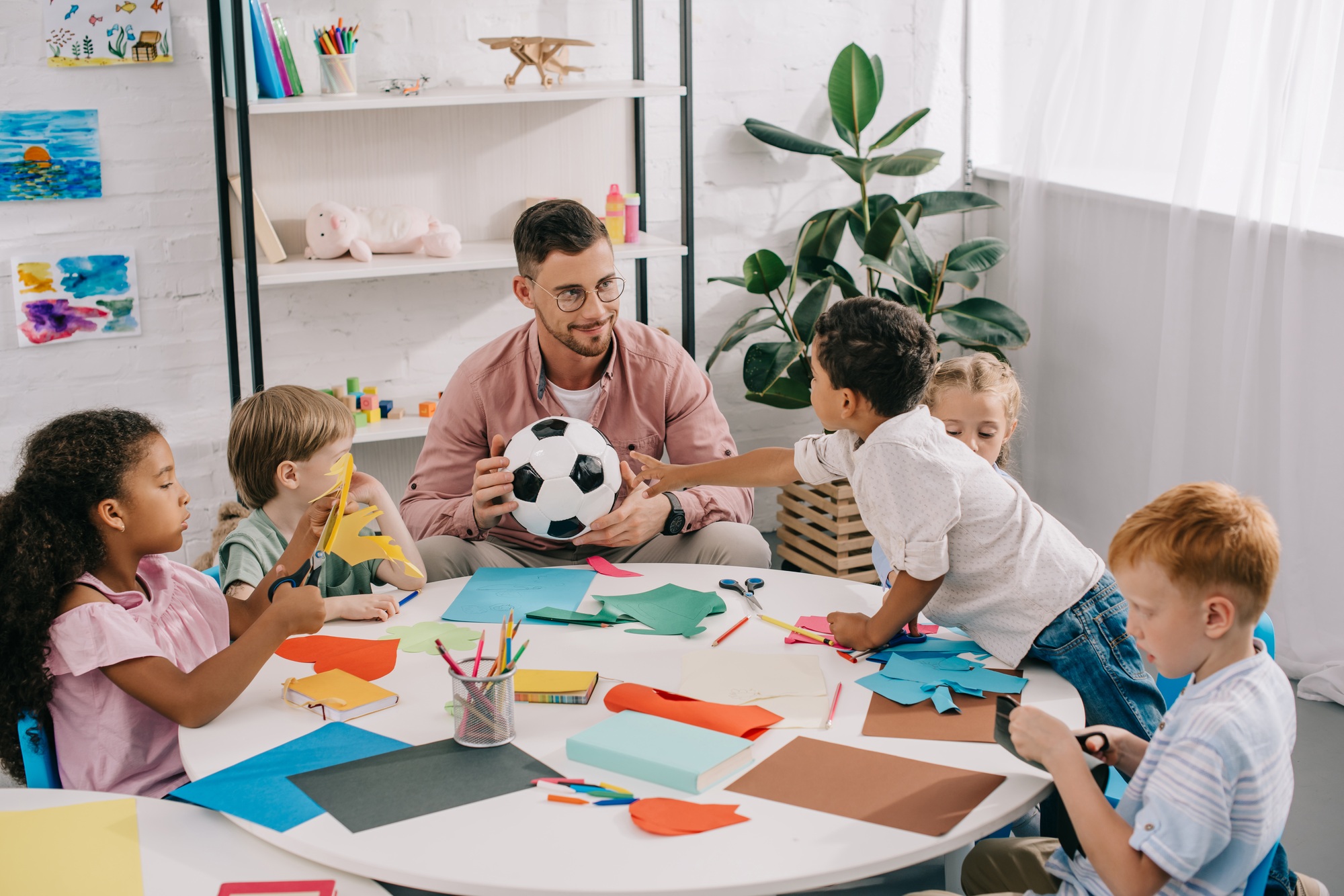 male teacher with soccer ball and multiracial preschoolers sitting at table with colorful papers in