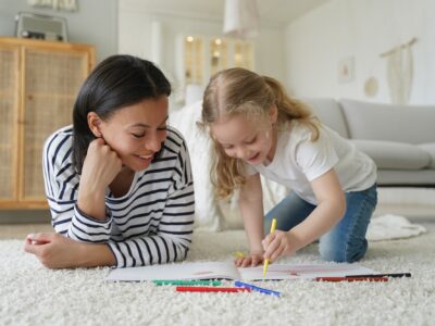 Mom, adopted child daughter draw with colorful pencils, lying on floor. Happy motherhood, adoption