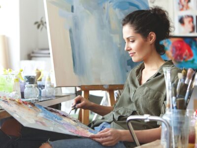 Portrait of beautiful female artist in casual clothes, mixing brightful colors, drawing on easel whi