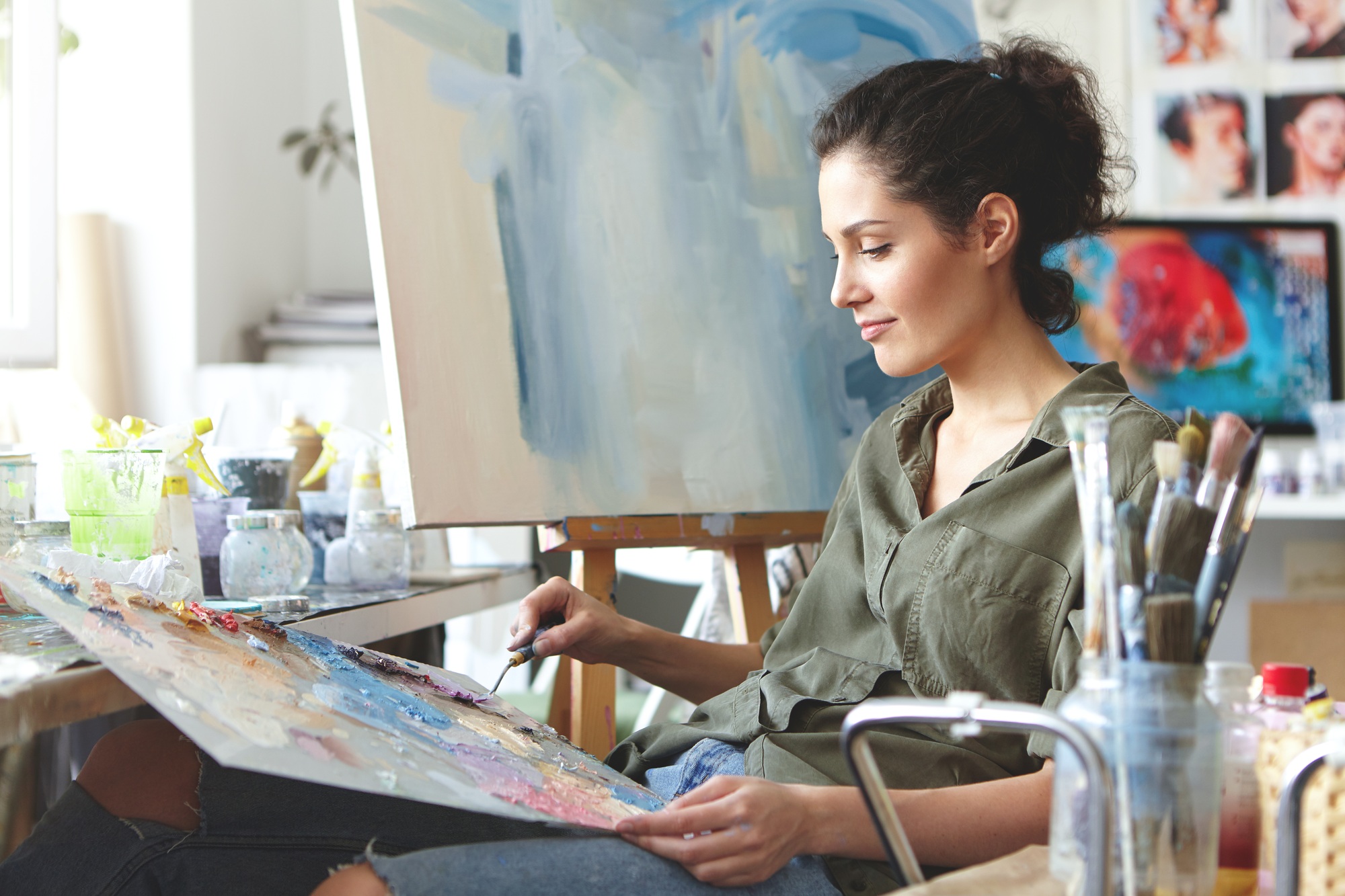 Portrait of beautiful female artist in casual clothes, mixing brightful colors, drawing on easel whi